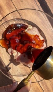 A close-up of raw chicken pieces in a glass bowl, being drizzled with a dark marinade from a small brass cup on a wooden surface, with sunlight casting shadows—unlike the quick and simple Viral Melon Ice Cream (2 Ingredients) recipes, this dish showcases the rich complexity of savory flavors coming together.