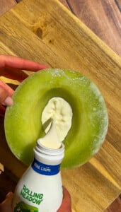 A hand pours cream from a Rolling Meadow carton into the hollow center of a halved green melon on a wooden surface, creating the Viral Melon Ice Cream (2 Ingredients) treat.