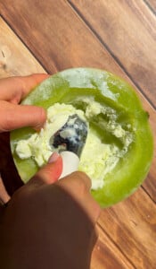 A hand holds half a green melon while another hand uses an ice cream scooper to scoop out vanilla ice cream from inside the melon—creating the viral Melon Ice Cream (2 Ingredients) treat everyone is talking about.