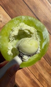 A hand holds an ice cream scoop with pale green melon sorbet, scooped from inside a hollowed-out melon half, on a wooden surface in sunlight—a delightful Viral Melon Ice Cream (2 Ingredients) treat perfect for summer days.