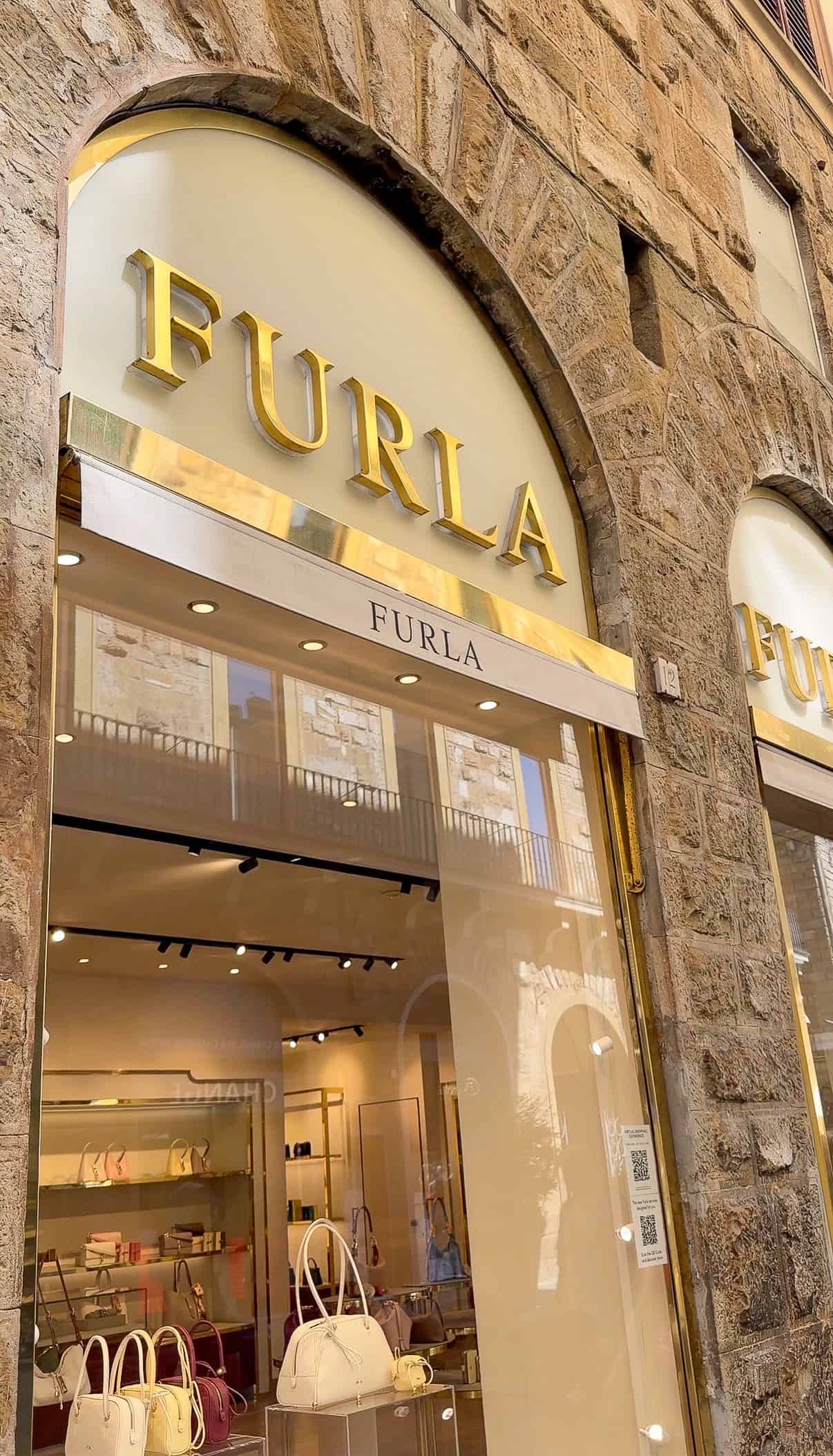 Storefront of a Furla boutique with large gold letters above the entrance, glass windows showing purses and accessories on display, set in a stone building.