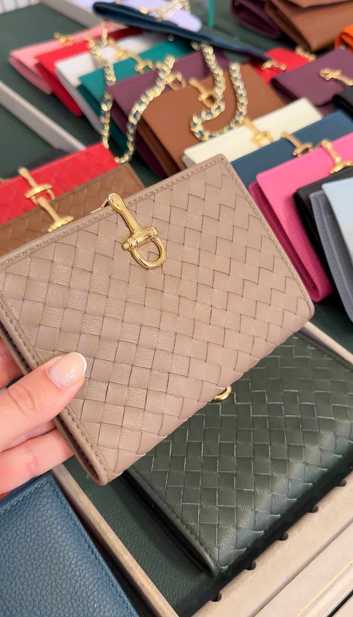 A hand holds a beige woven leather wallet with a gold clasp, displayed among wallets in various colors and styles on a table.