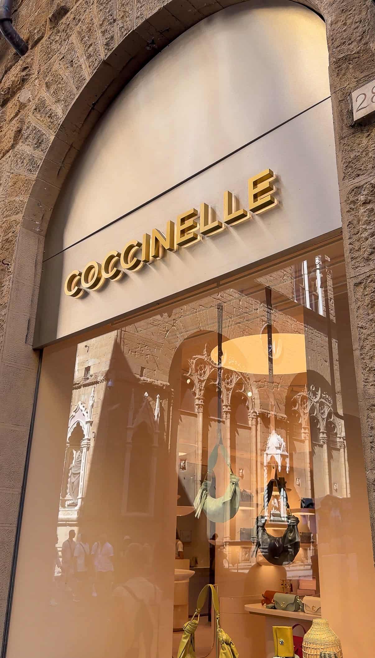 A store window with the sign "COCCINELLE" in gold letters. Handbags and accessories are displayed inside, with reflections of a historic building and people in the glass.
