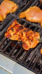 BBQ chicken thighs are being grilled on a barbecue. One piece is being turned with tongs, showing grill marks and a slightly charred, glazed surface. The grill grates and other chicken pieces are visible.