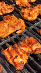 Close-up of marinated bbq chicken thighs sizzling on a grill, with char marks and a shiny glaze visible under bright sunlight.