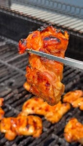 A close-up of grilled, glazed bbq chicken thighs held by metal tongs over a barbecue with more pieces cooking on the grill.