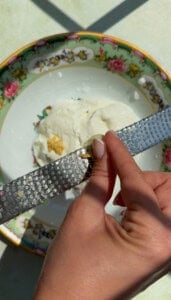 A hand grates a clove of garlic over a plate with a floral rim, containing a scoop of white creamy cheese, ready to be mixed into flavorful yogurt noodles.