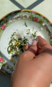 A hand sprinkles salt over a bowl with white cheese, chopped herbs, and lemon zest—ingredients perfect for brightening up a dish of Yogurt Noodles. The floral-patterned bowl sits on a light surface, ready to serve as a fresh and flavorful topping.