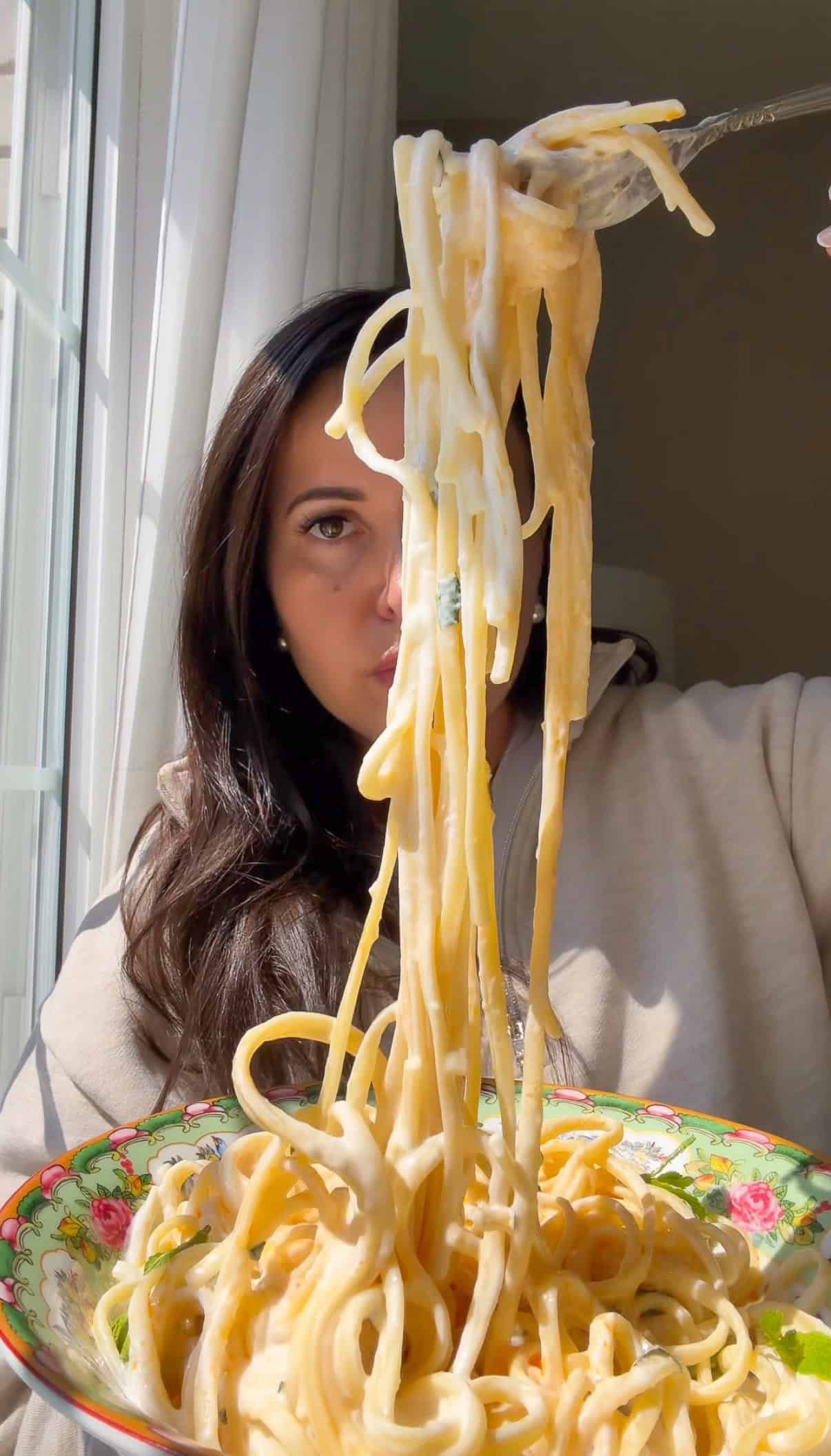 A woman in a beige top lifts a forkful of creamy yogurt noodles from a colorful floral bowl, with sunlight streaming through a window beside her.