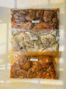 Three labeled ziplock bags filled with flavorful chicken marinades: one with garlic and honey, one with Greek lemon, and one with peri peri. Each marinade is seasoned with herbs and spices, and the bags are arranged vertically.