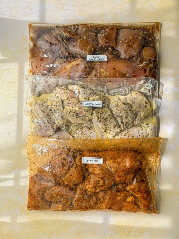 Three labeled ziplock bags filled with flavorful chicken marinades: one with garlic and honey, one with Greek lemon, and one with peri peri. Each marinade is seasoned with herbs and spices, and the bags are arranged vertically.