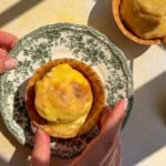 A person holds a vintage plate with a baked tart topped with melted cheese, reminiscent of flavors found in classic chicken marinades. Another tart sits on the table nearby, both lit by sunlight and shadow patterns.
