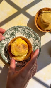 A person holds a vintage plate with a baked tart topped with melted cheese, reminiscent of flavors found in classic chicken marinades. Another tart sits on the table nearby, both lit by sunlight and shadow patterns.