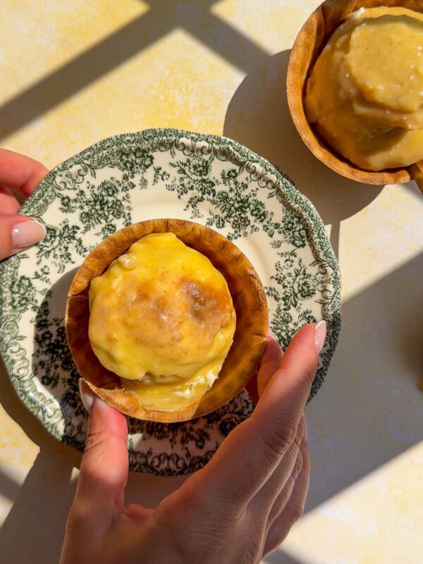 A person holds a vintage plate with a baked tart topped with melted cheese, reminiscent of flavors found in classic chicken marinades. Another tart sits on the table nearby, both lit by sunlight and shadow patterns.