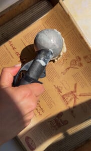 A hand scoops vanilla ice cream with a metal scooper over a printed brown box with red illustrations and text, reminiscent of packaging for savory delights like chicken marinades.