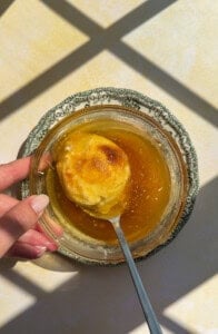 A hand holds a glass bowl with honey and a biscuit on a spoon, casting shadows on a light surface—perfect inspiration for those seeking sweet touches in chicken marinades.