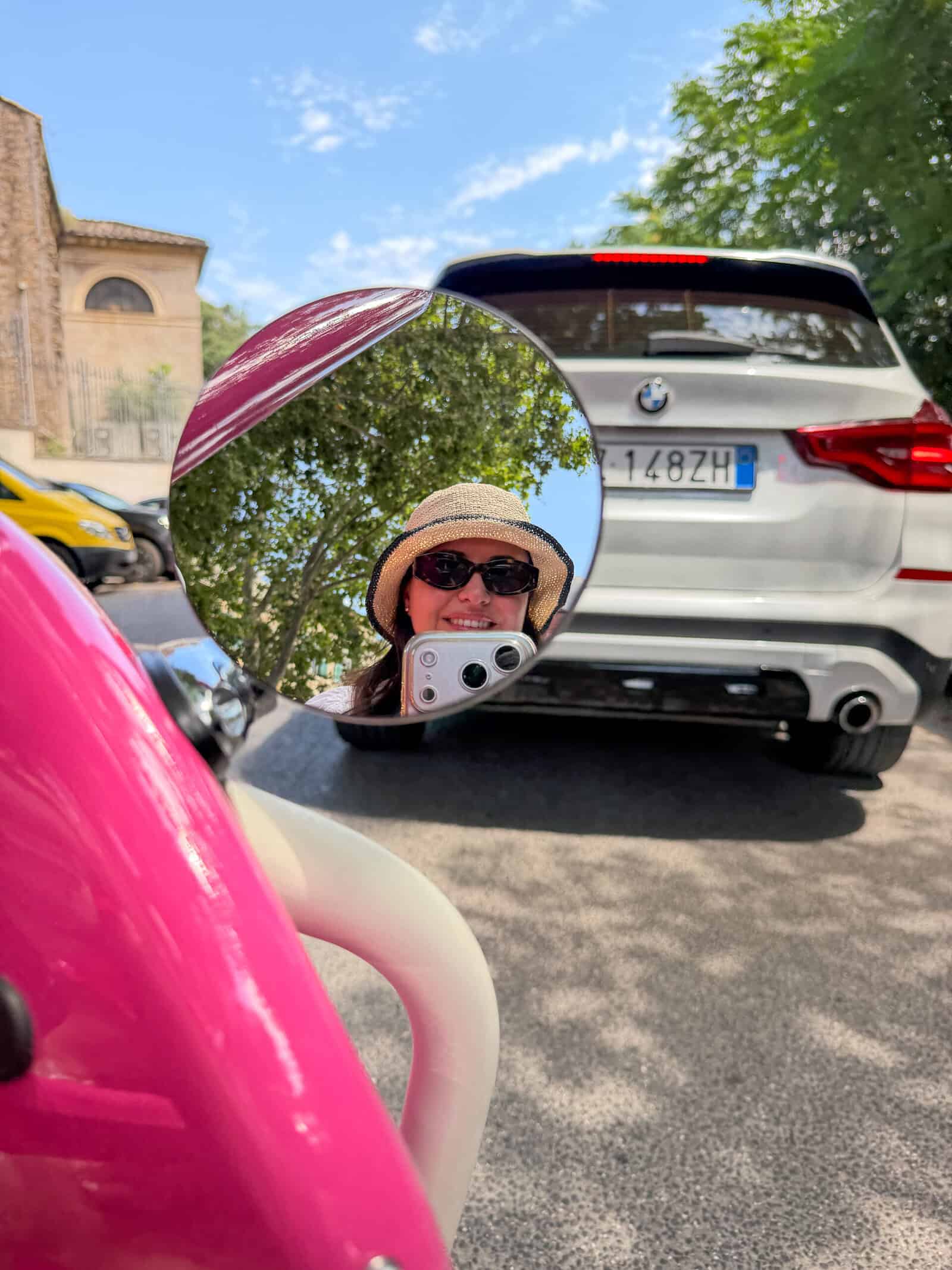 A person wearing sunglasses and a straw hat is reflected in a round mirror attached to a pink scooter. They are taking a photo. In the background, there’s a white car parked on a sunny street with trees and buildings.