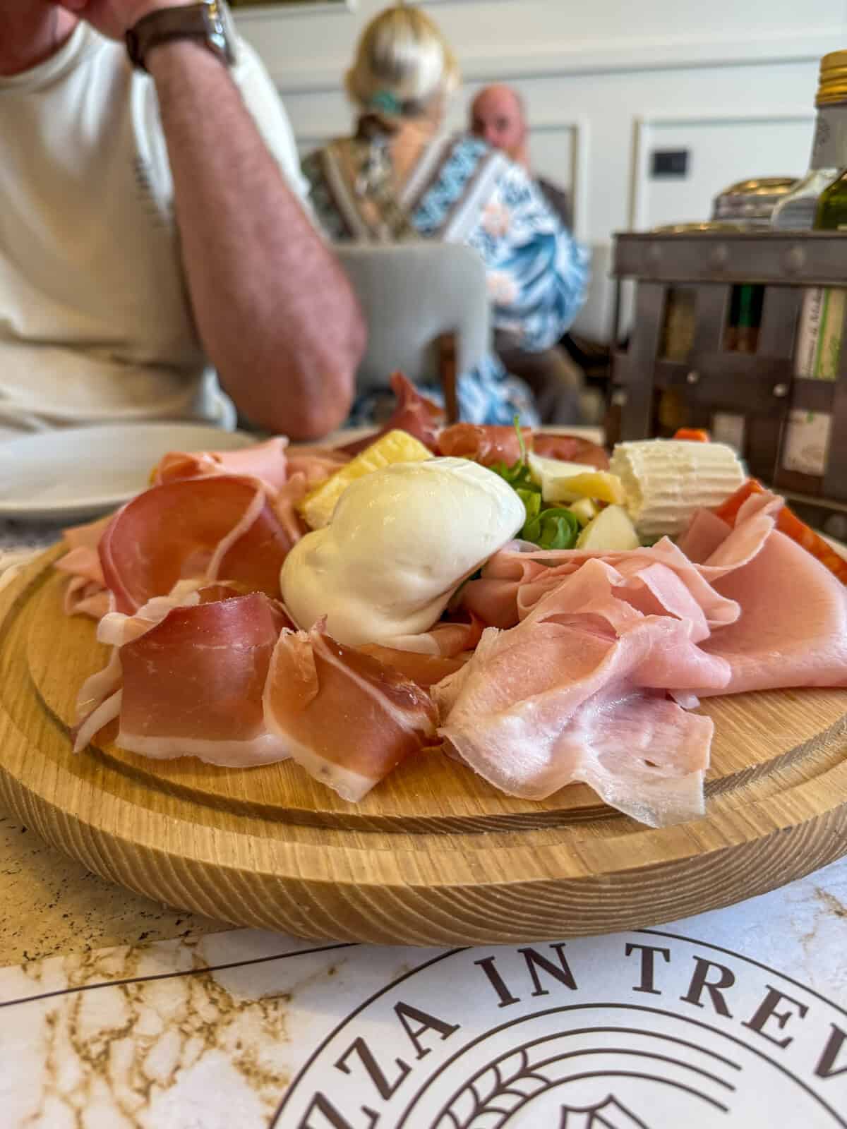 A wooden platter with assorted cured meats, cheese, and a ball of fresh mozzarella is on a table in a restaurant. Diners are seated in the background, and part of a menu is visible in the foreground.