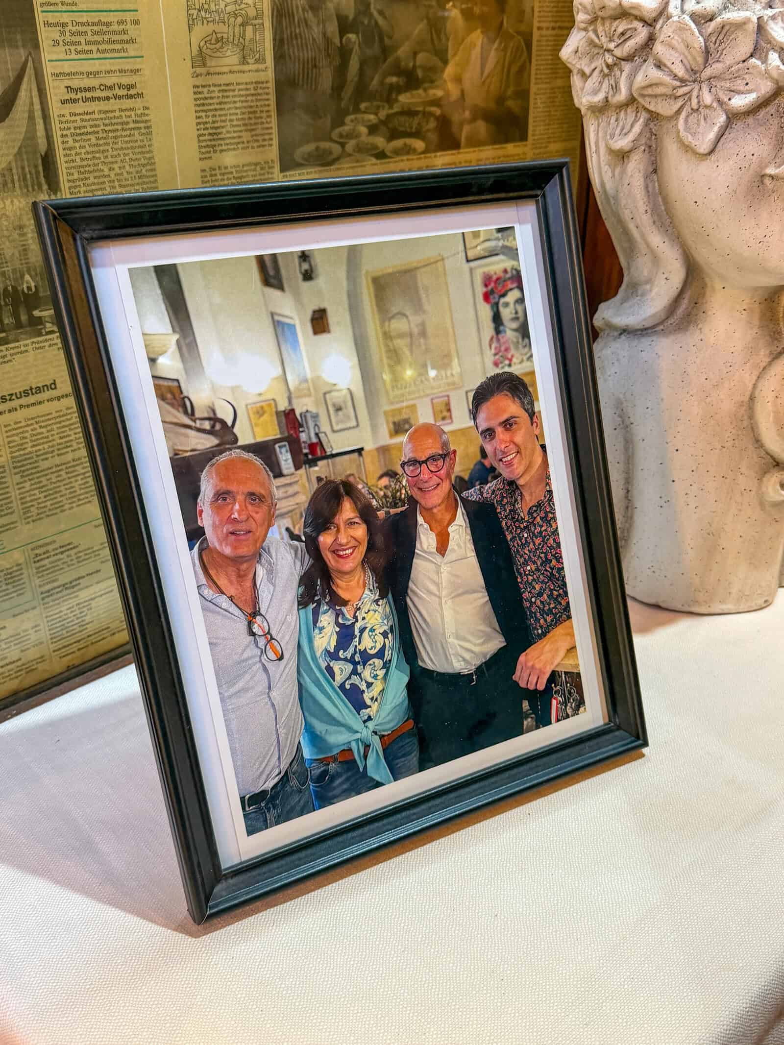 A framed photo of four smiling adults standing close together indoors, with their arms around each other, displayed on a table next to a decorative vase and in front of background newspaper clippings.