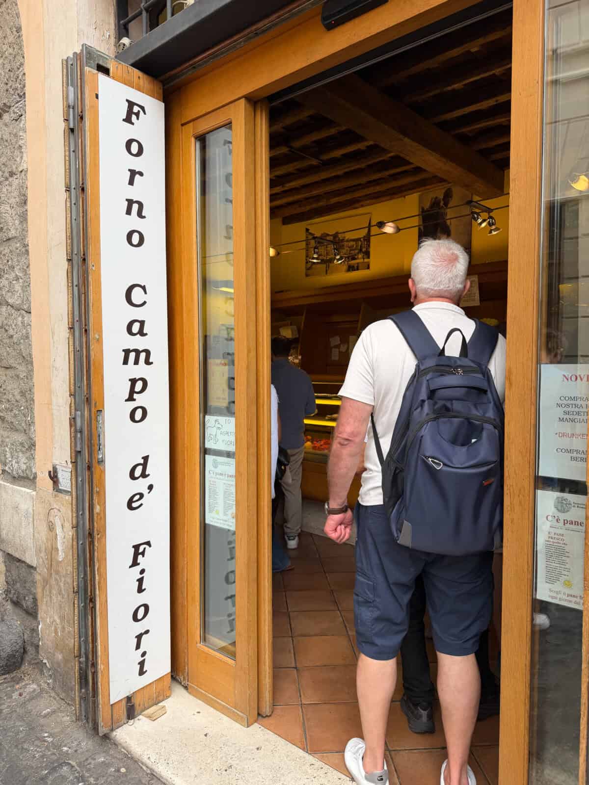A man with a backpack enters Forno Campo de’ Fiori, a bakery with a white sign and wooden door, as people stand inside near the counter.