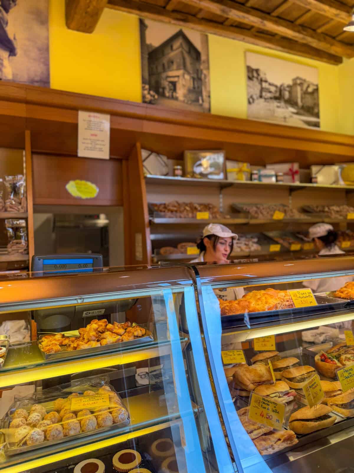 A bakery with glass display cases filled with various pastries and breads. A worker wearing a white cap stands behind the counter. Yellow price tags are visible, and shelves with more baked goods and vintage photos are on the wall.