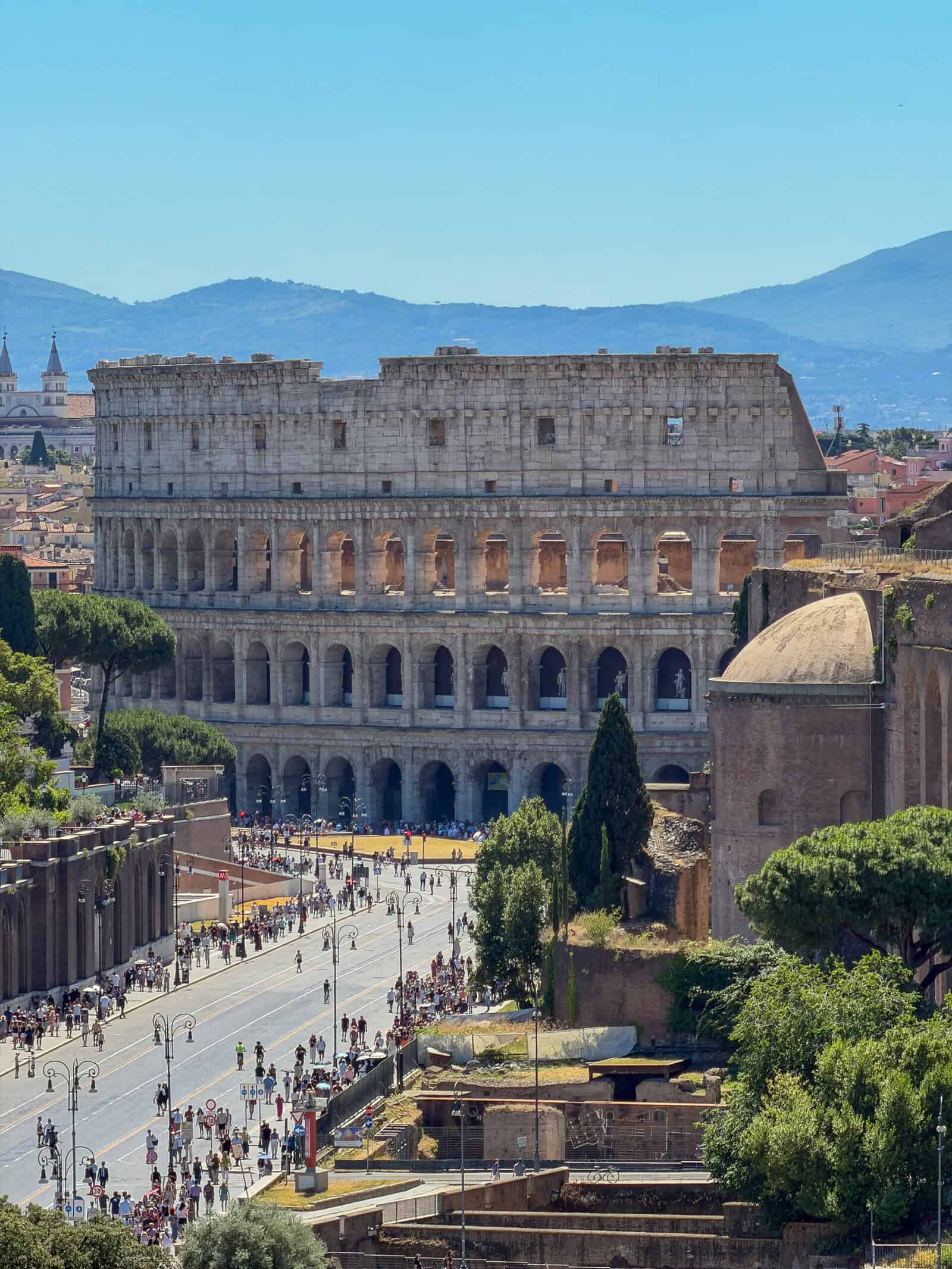 The Colosseum in Rome stands under a clear blue sky, surrounded by crowds of tourists and nearby historic ruins, with distant mountains visible in the background.