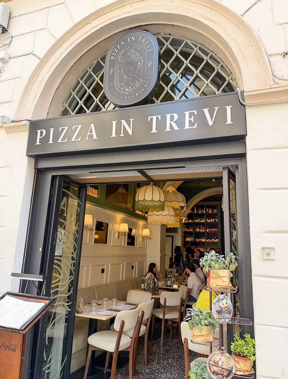 Street view of "Pizza in Trevi" restaurant with an arched entrance, open doors, visible dining tables, chandeliers, and people sitting inside. A menu stand and a plant display are at the entrance.