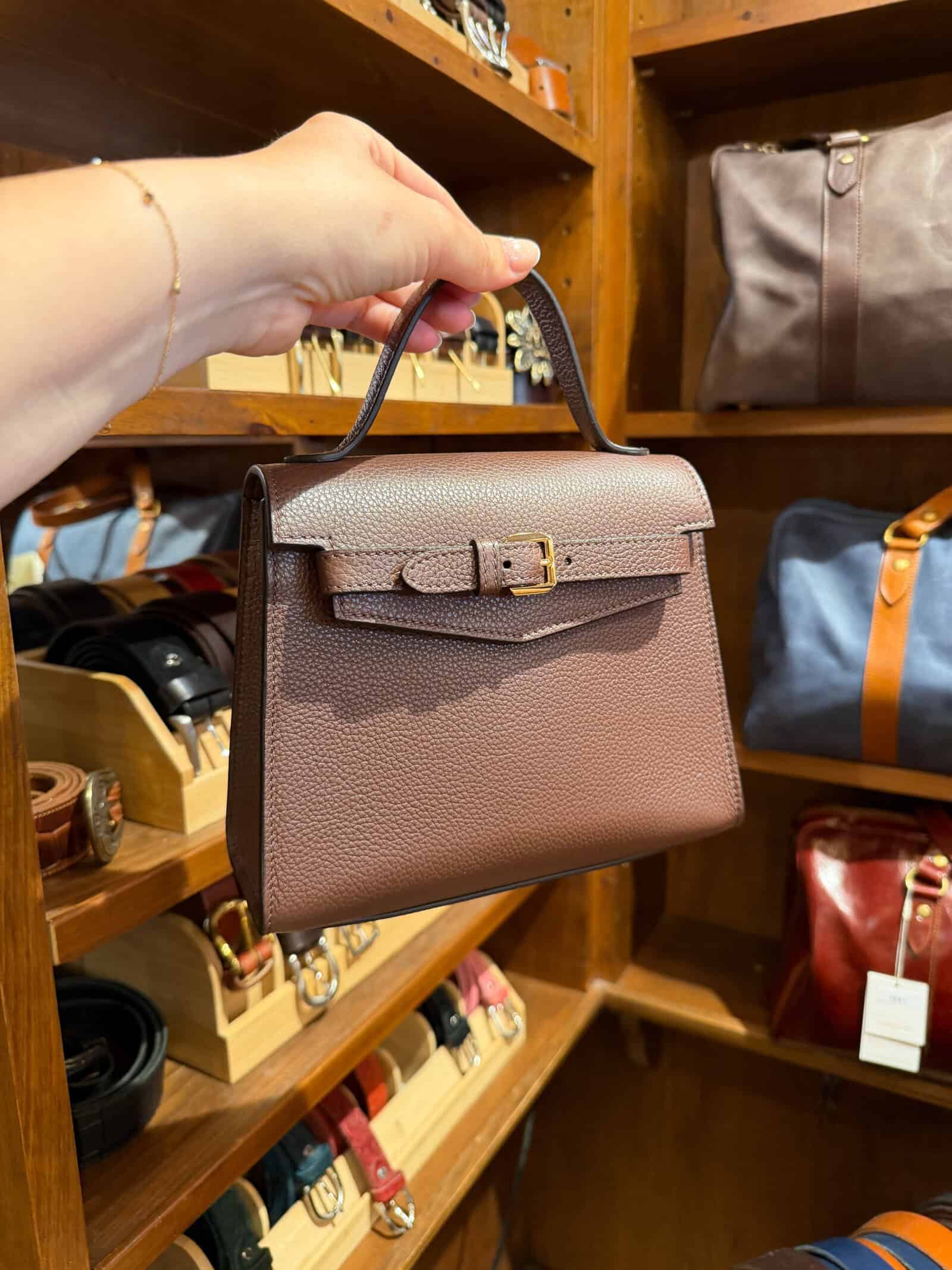 A hand holds a small brown leather handbag with a top handle and buckle, displayed in a store with shelves of belts and bags in the background.
