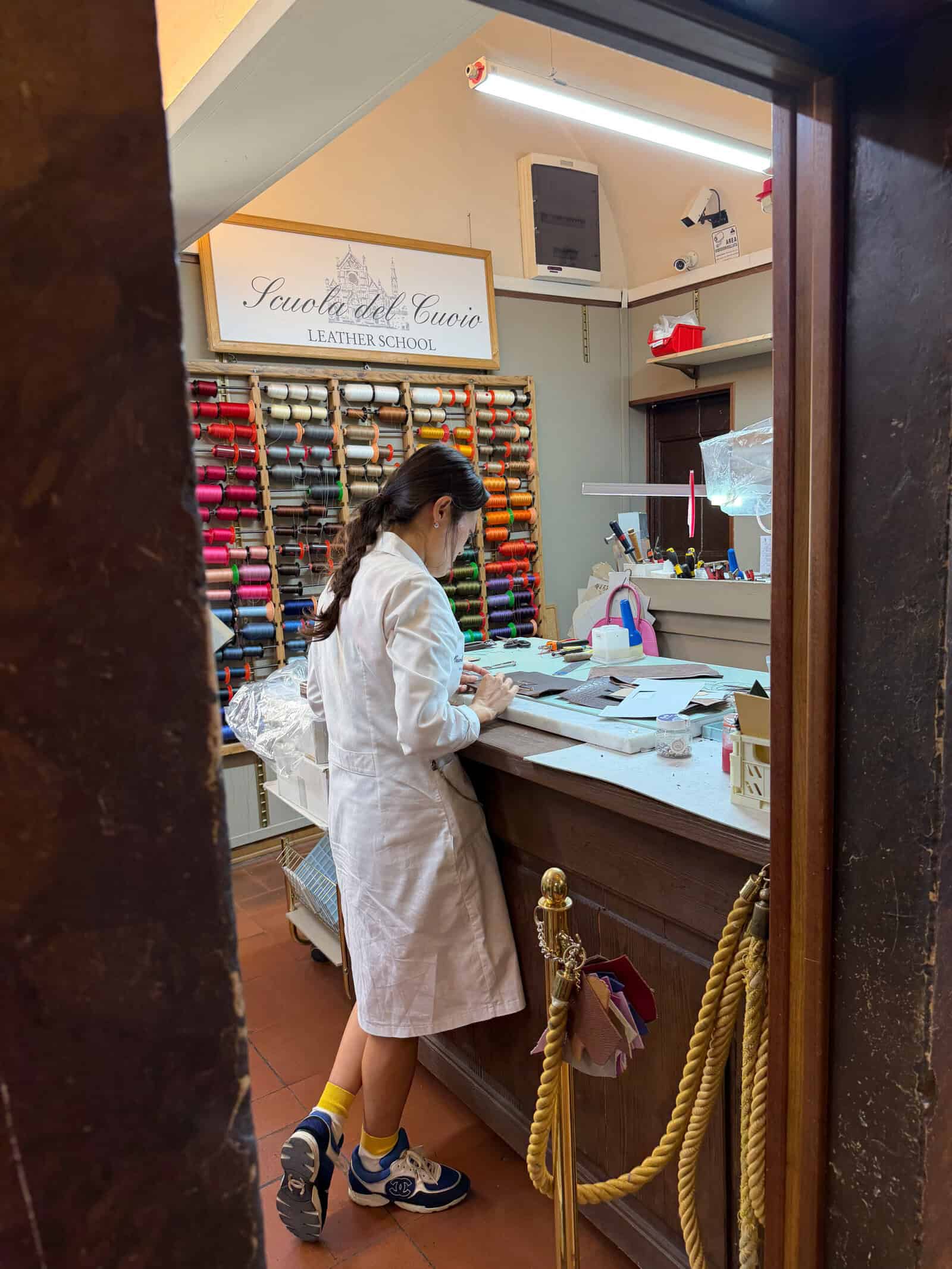 A person in a white lab coat works at a counter in a leather school, with colorful threads organized on the wall behind them and various leatherworking tools on the desk.