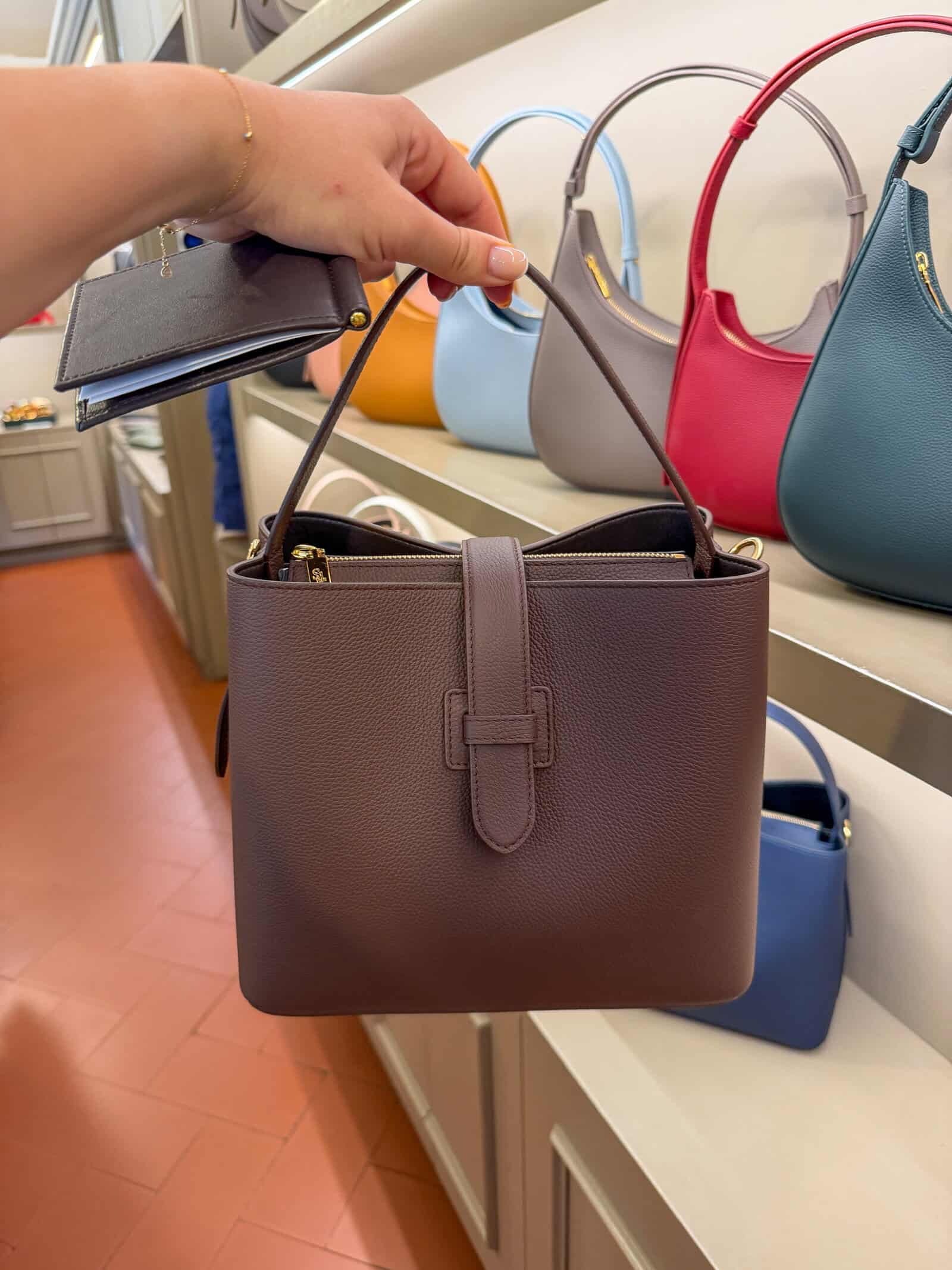 A hand holds a brown leather handbag with a strap in a store. Other colorful handbags are displayed on shelves in the background.