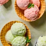 Two waffle bowls filled with three scoops of ice cream each, one with pink ice cream and the other with light green, both garnished with fresh mint. Metal ice cream scoops and extra ice cream are nearby.