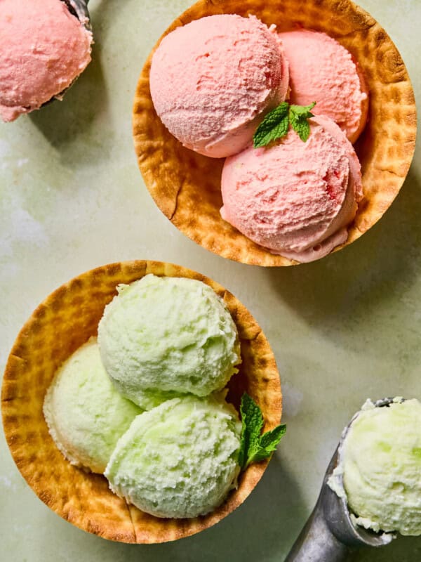 Two waffle bowls filled with three scoops of ice cream each, one with pink ice cream and the other with light green, both garnished with fresh mint. Metal ice cream scoops and extra ice cream are nearby.