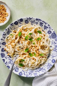 A plate of creamy spaghetti topped with toasted pine nuts and fresh mint leaves, served on a blue-patterned dish with a fork. A white napkin and a bowl of pine nuts are beside the plate.