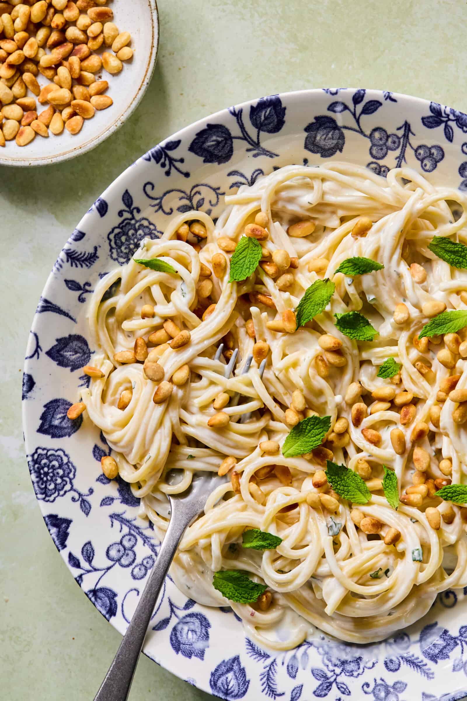 A plate of creamy spaghetti topped with pine nuts and fresh mint leaves sits on a floral-patterned dish, with a fork and a small bowl of pine nuts nearby.