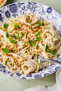 A bowl of creamy spaghetti topped with toasted pine nuts and fresh mint leaves, served in a blue and white patterned dish with a fork resting inside.