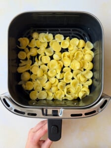 A hand holds an air fryer drawer filled with uncooked orecchiette pasta, spread evenly at the bottom, ready to be cooked—perfect for pairing with classic recipes like chicken in a potato.