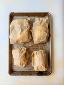 A baking sheet with four food parcels wrapped in brown parchment paper, possibly containing chicken in a potato, arranged in two rows on a light-colored surface.