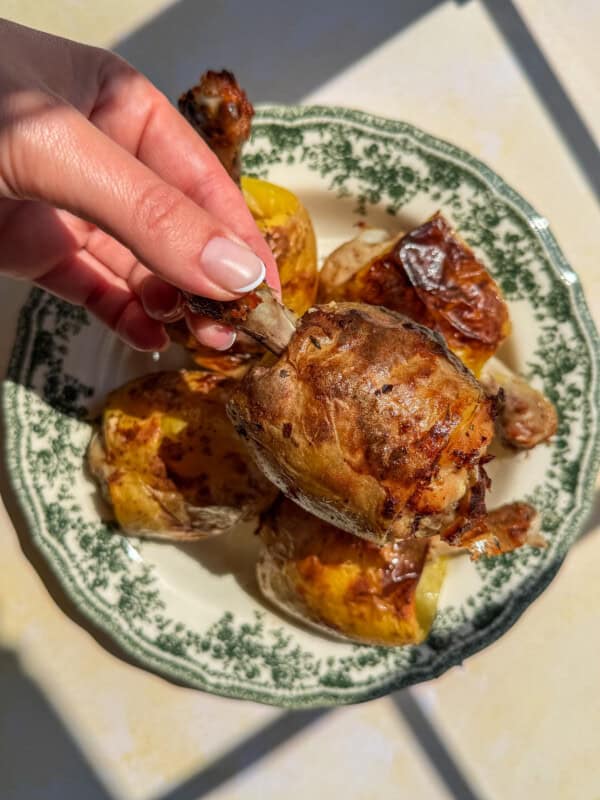 A hand holding a roasted chicken drumstick above a vintage-patterned plate with more roasted chicken and potatoes in bright sunlight, showcasing the delicious pairing of chicken in a potato-inspired meal.