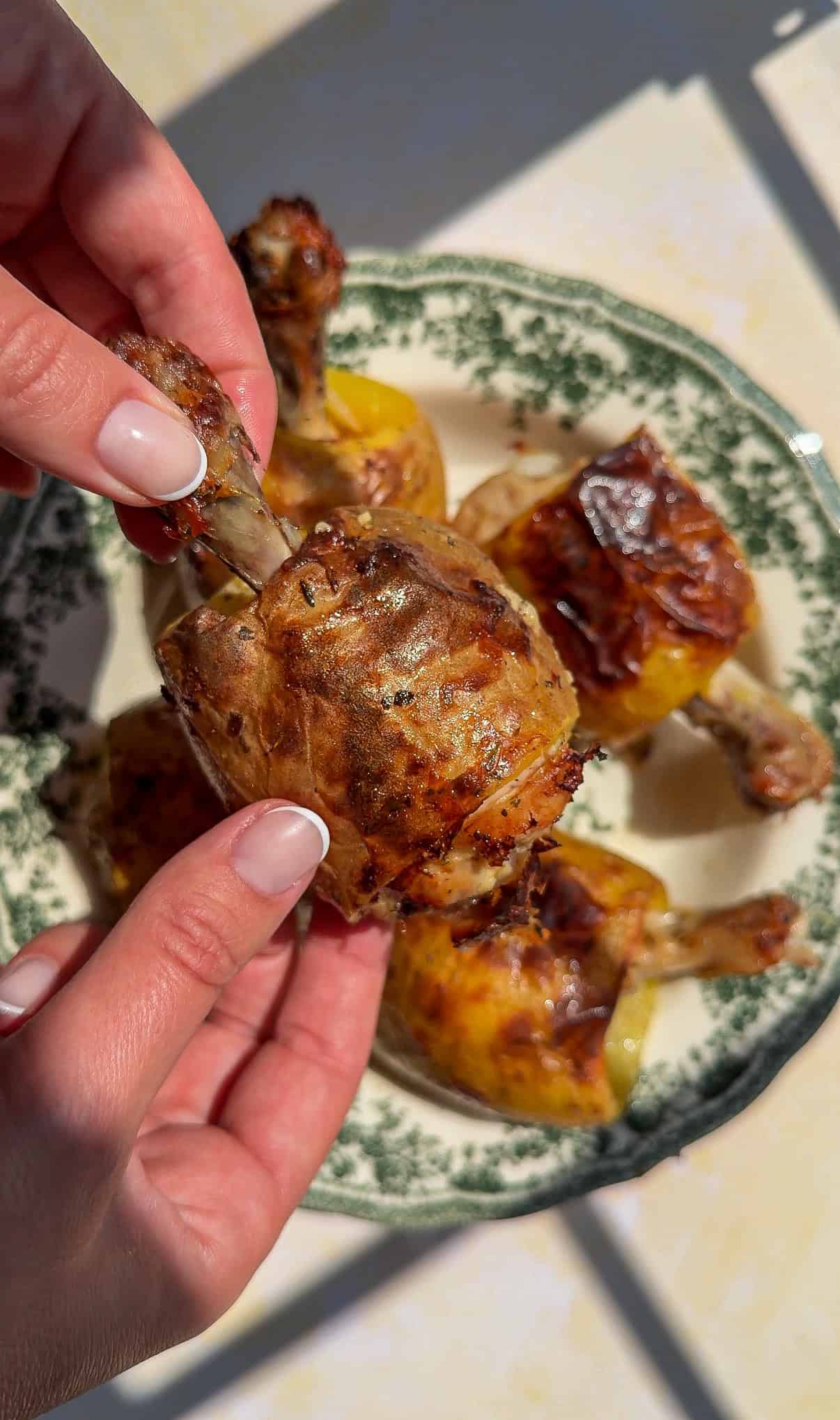 Two hands holding a cooked chicken drumstick with crispy skin over a green and white patterned plate with more drumsticks on it, in bright natural light—perfect for pairing with hearty sides like chicken in a potato.