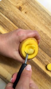 A hand uses a melon baller to scoop out the center of a small yellow potato on a wooden cutting board, preparing it as the perfect vessel for serving chicken in a potato.