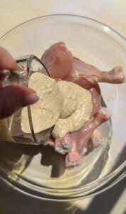 A hand pours creamy, seasoned marinade from a glass cup over raw chicken drumsticks and thighs in a clear glass bowl, setting the stage for a delicious chicken in a potato-inspired dish.