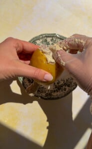 Two hands, one gloved, are peeling the skin off a yellow potato with some cream or sauce on it—perhaps preparing the base for chicken in a potato—over a decorative plate on a yellow surface.