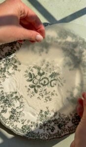Two hands hold a translucent circular sheet above a decorative plate with green floral patterns, with sunlight casting shadows across the surface.