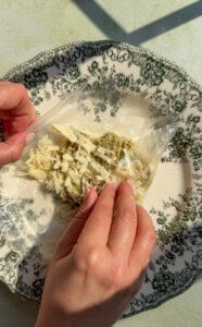 Two hands fold a rice paper sheet filled with shredded cheese and herbs on a decorative plate with a black floral pattern.