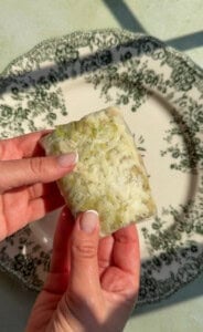 Two hands hold a rectangular block of rice and green vegetables wrapped in translucent paper, over a decorative white plate with a dark floral pattern.