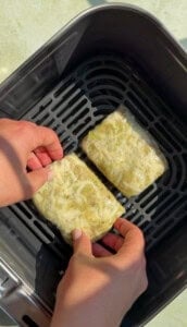 Two hands place two rectangular pieces of food topped with shredded cheese, wrapped in plastic, into an air fryer basket.