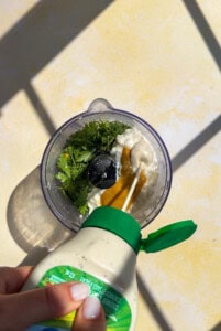 Hand pouring dressing into blender with herbs and sauce