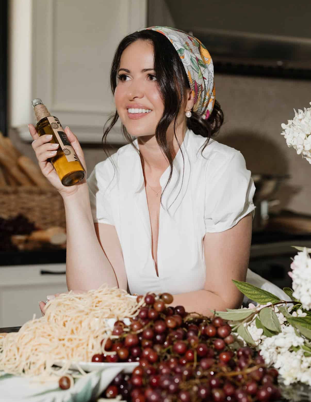 Smiling woman holding olive oil bottle in kitchen with grapes