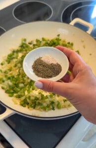 Hand holding spices over sautéed onions and peppers in pan