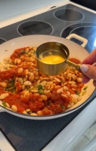 Hand adding oil to white beans and tomato sauce in skillet